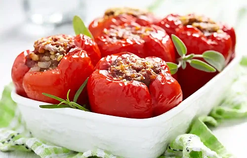 Baltame keraminiame kepimo inde patiektos iškeptos, ryškiai raudonos saldžiosios paprikos, gausiai įdarytos mėsos faršu ir papuoštos šviežiais šalavijų bei rozmarinų lapeliais.